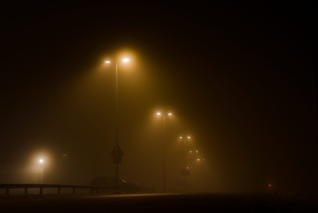 Parkplatz bei Nacht mit mehreren leuchtenden Masten, Fahrzeugen und leichtem Nebel.