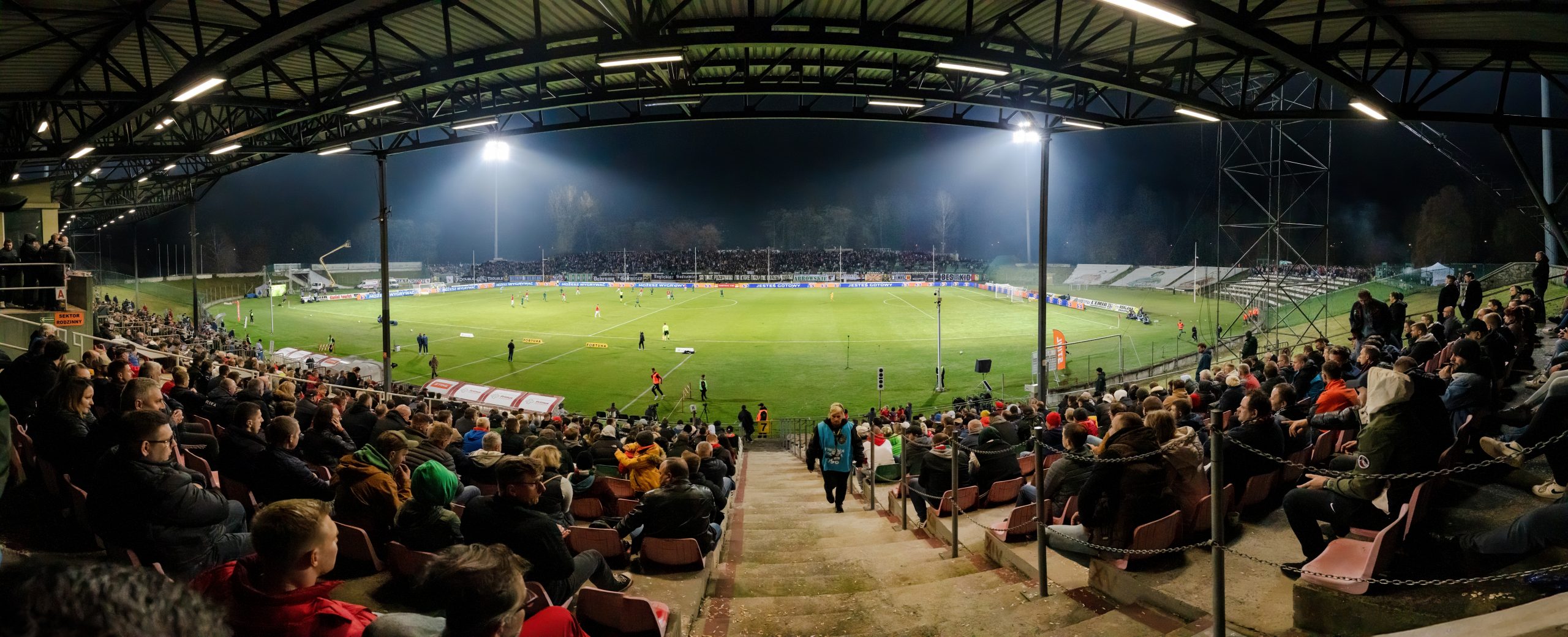 Fußballstadion bei Nacht mit hell ausgeleuchtetem Spielfeld, Tribünen und Flutlichtanlage.
