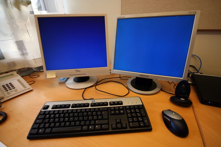 Büroschreibtisch mit Bildschirm, Tastatur und Arbeitsplatzleuchte vor einer hellen Wand.