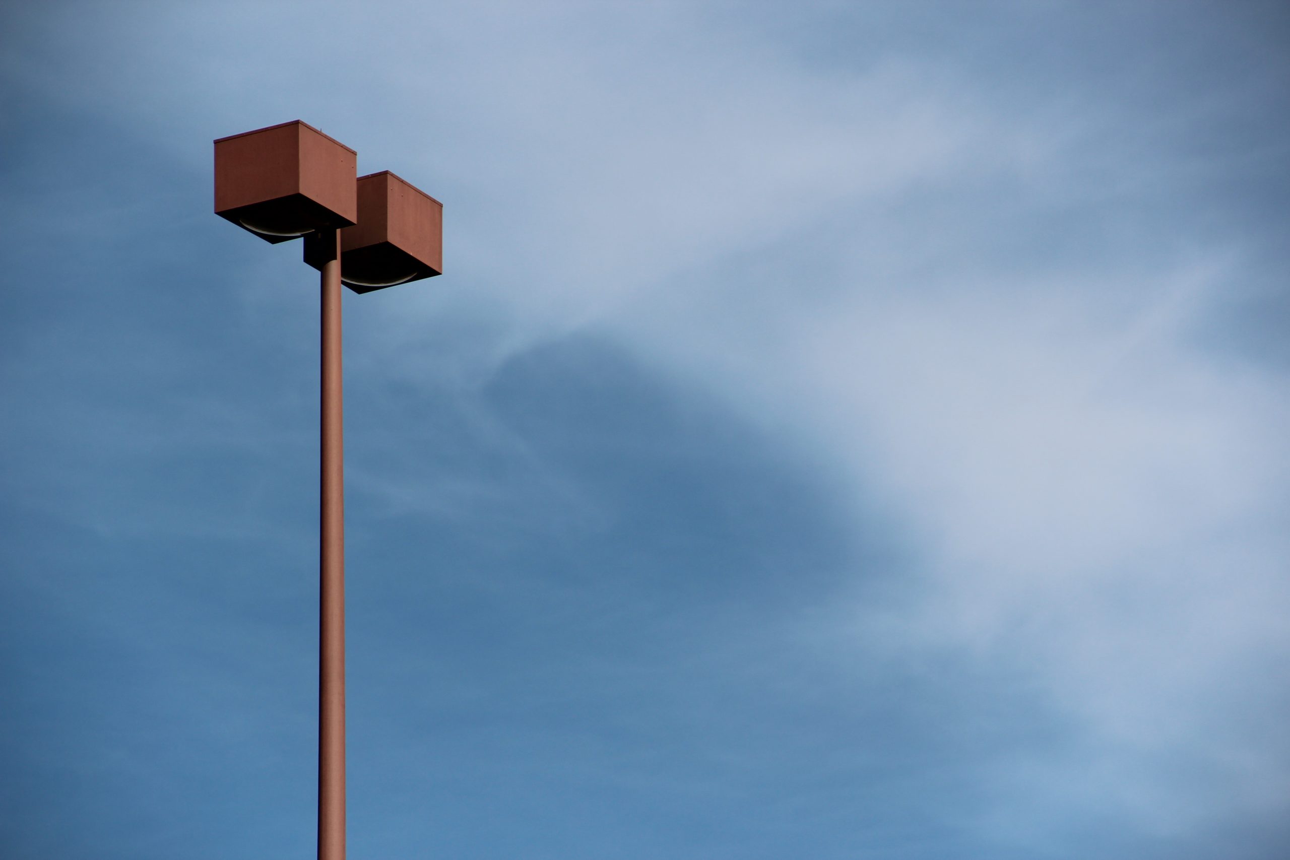 Hoher Parkplatz-Lichtmast mit zwei Leuchten vor blauem Himmel und Bäumen.