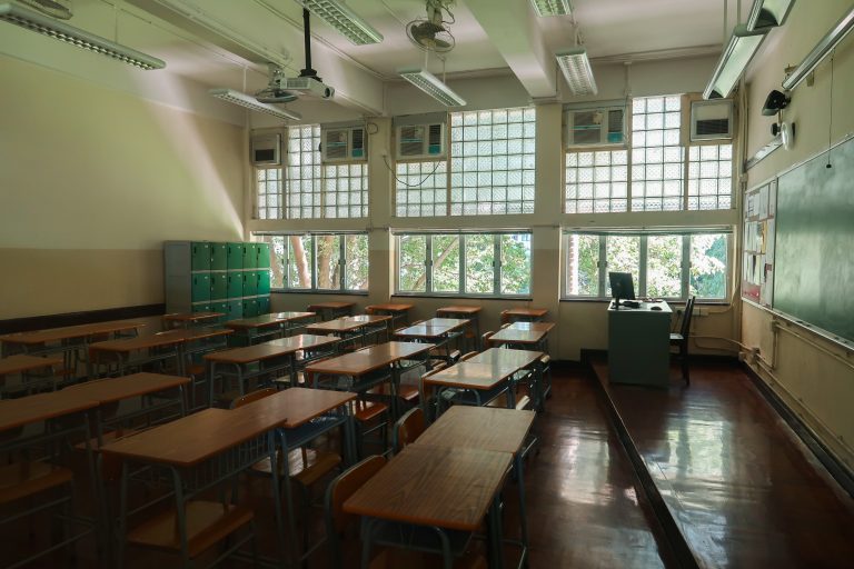 Klassenzimmer mit Tischreihen, Stühlen, Tafel und mehreren Deckenleuchten.