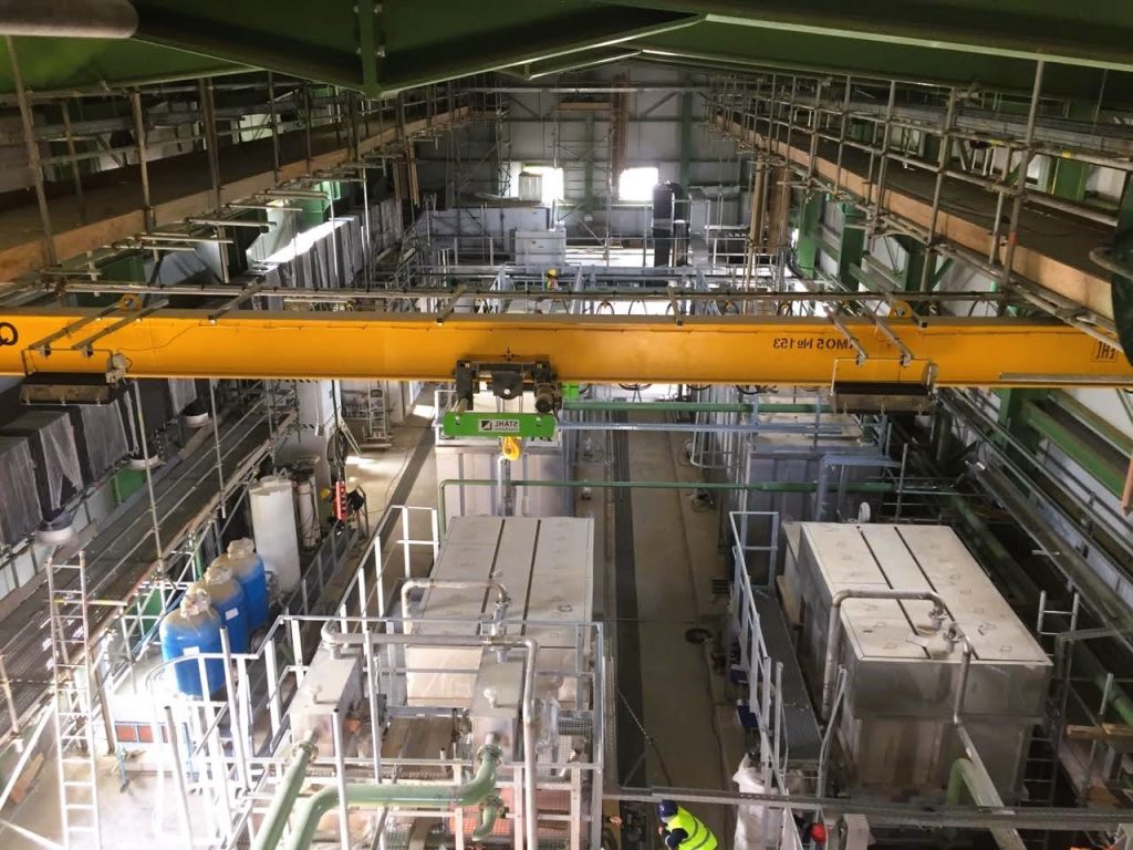 Blick in eine aktive industrielle Produktionsanlage mit Kranbahn, Maschinen und Deckenbeleuchtung.